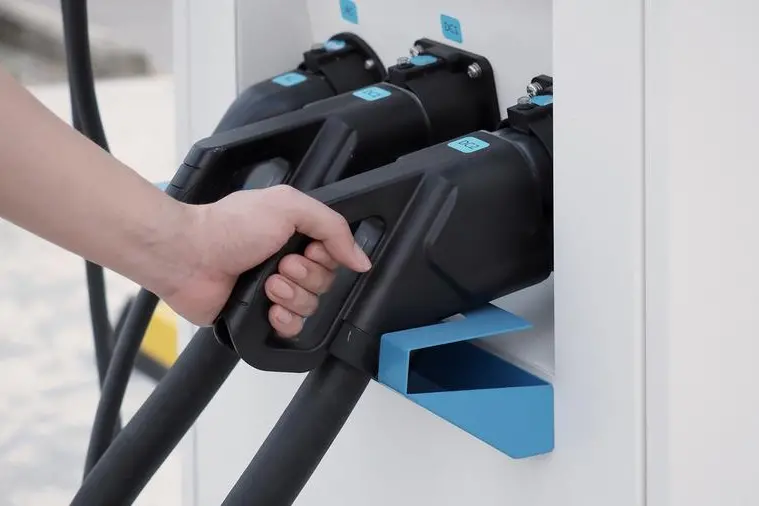 Hand holding EV charging plug in front of camera with blurred background of outdoor. Driver of electric car hold batteries charger plug. Sustainable energy powered electric charging station background. Image used for illustrative purpose. Image courtesy: Getty Images/ PpengCreative , Getty Images